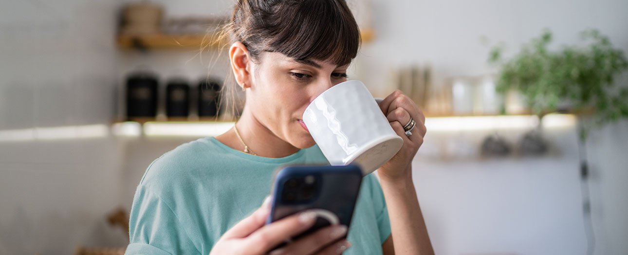 Frau trinkt eine Tasse Kaffee und blickt auf Ihr mobiles Telefon