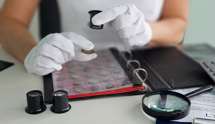Person mit weißen Handschuhen betrachtet eine Münze unter der Lupe