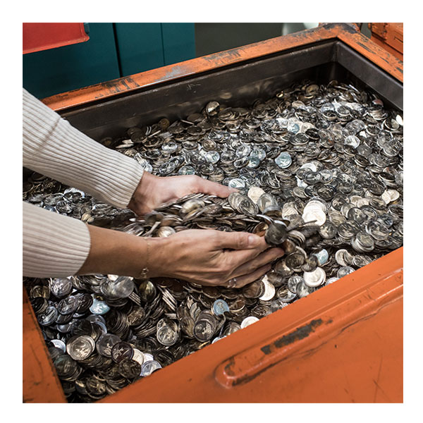 Zwei Hände greifen in einen orangefarbenen Container voller 2-Euro- Münzen.