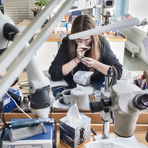 Eine Stempelschleiferin betrachtet einen Münzstempel durch die Lupe. Im Vordergrund sind diverse Werkzeuge aus der Werkstatt zu sehen.