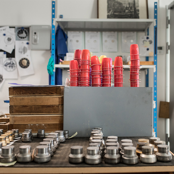 Im Vordergrund liegen Prägestempel auf einem Tisch. Dahinter stehen gestapelte Joghurtbecher auf einer Anrichte.