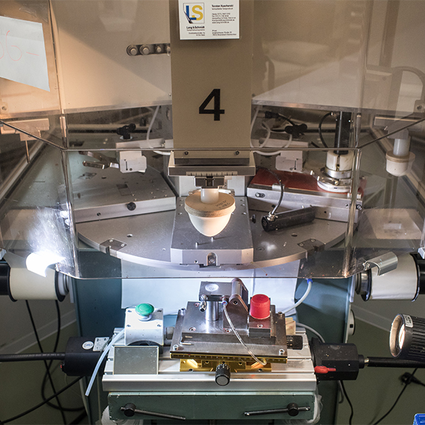 Eine Stempelschleiferin betrachtet einen Münzstempel durch die Lupe. Im Vordergrund sind diverse Werkzeuge aus der Werkstatt zu sehen.